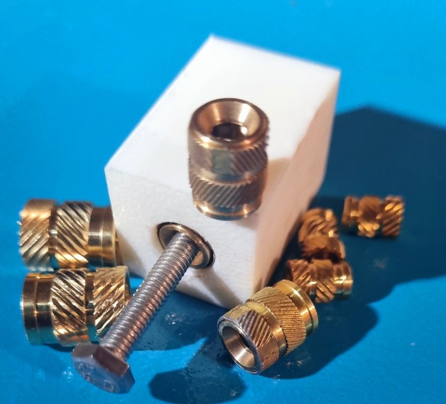 Boccole BLOWLOCK in ottone per plastica accanto a un blocco campione bianco con vite